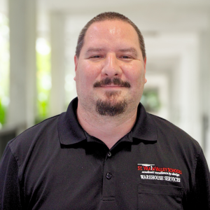 A man in a black shirt with "St. Vrain Valley Schools Warehouse Services" stands smiling in a bright, blurred background.