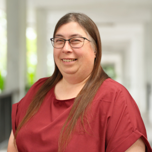 A woman with long brown hair and glasses smiles, wearing a maroon shirt in a bright, blurred indoor setting.