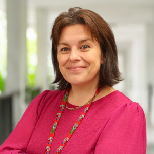 Smiling woman in a pink blouse with a colorful necklace, standing in a bright, modern indoor space.
