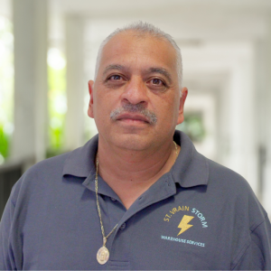A man with gray hair and a mustache, wearing a blue shirt with a logo, stands in a well-lit corridor.