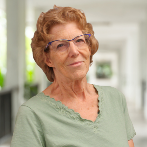 A smiling older woman with curly hair, wearing glasses and a green shirt, stands in a bright corridor.