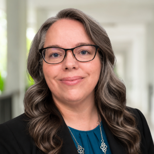 A smiling woman with long, wavy hair and glasses, wearing a black blazer and teal top, stands in a bright corridor.