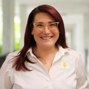 A woman with red hair and glasses smiles in a bright hallway, wearing a white shirt with a logo for Nutrition Services.