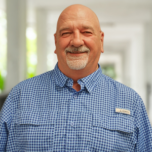 Smiling man with a bald head and goatee, wearing a blue checkered shirt, standing in a bright, blurred interior.