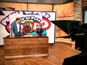 A male and female news anchor on a TV set. They're standing behind a tall table on the left. 7th grader Wolf Danzer is to their right demonstrating how his game words during a live news broadcast.