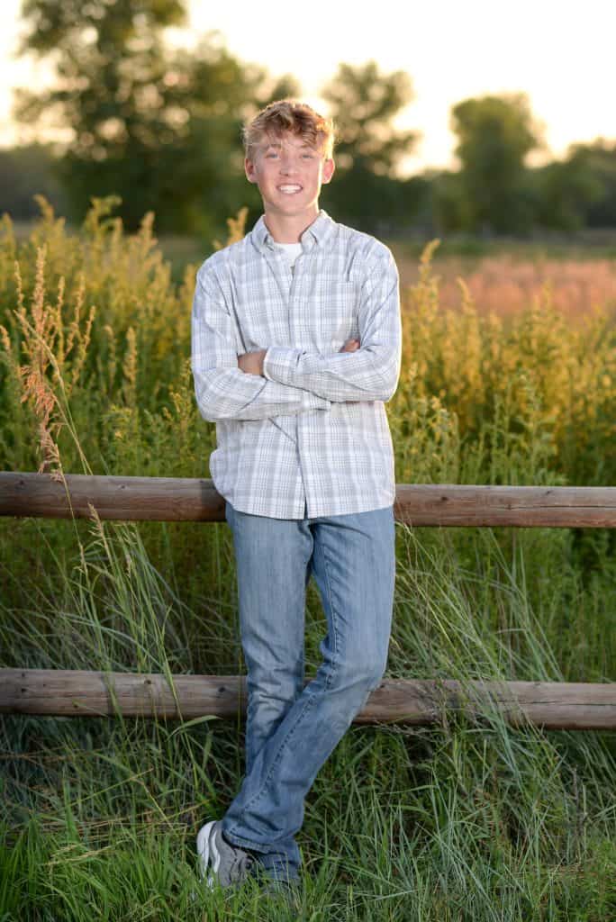 Aiden Phelan, Skyline High School graduate leaning on a fence in a grassy area and smiling