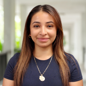 A smiling woman with long brown hair, wearing a navy shirt and gold necklace, stands in a bright, blurred indoor setting.