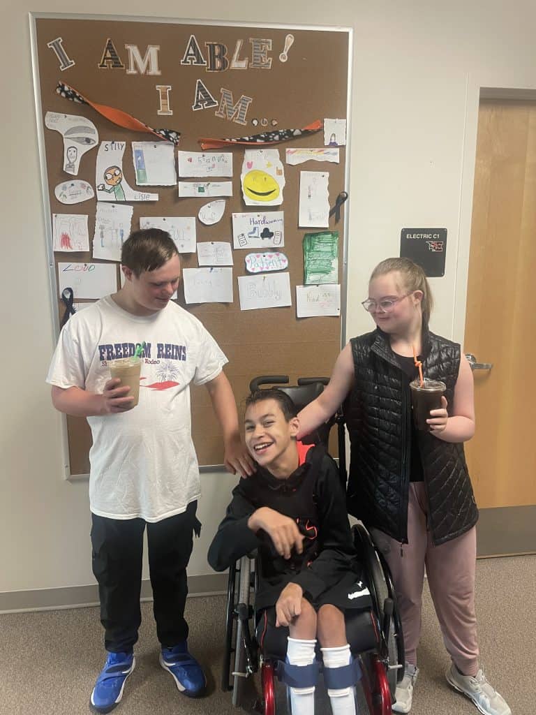 3 students pose with coffee cups. 2 students are looking down with their hands on the shoulder of a student in a wheel chair smiling