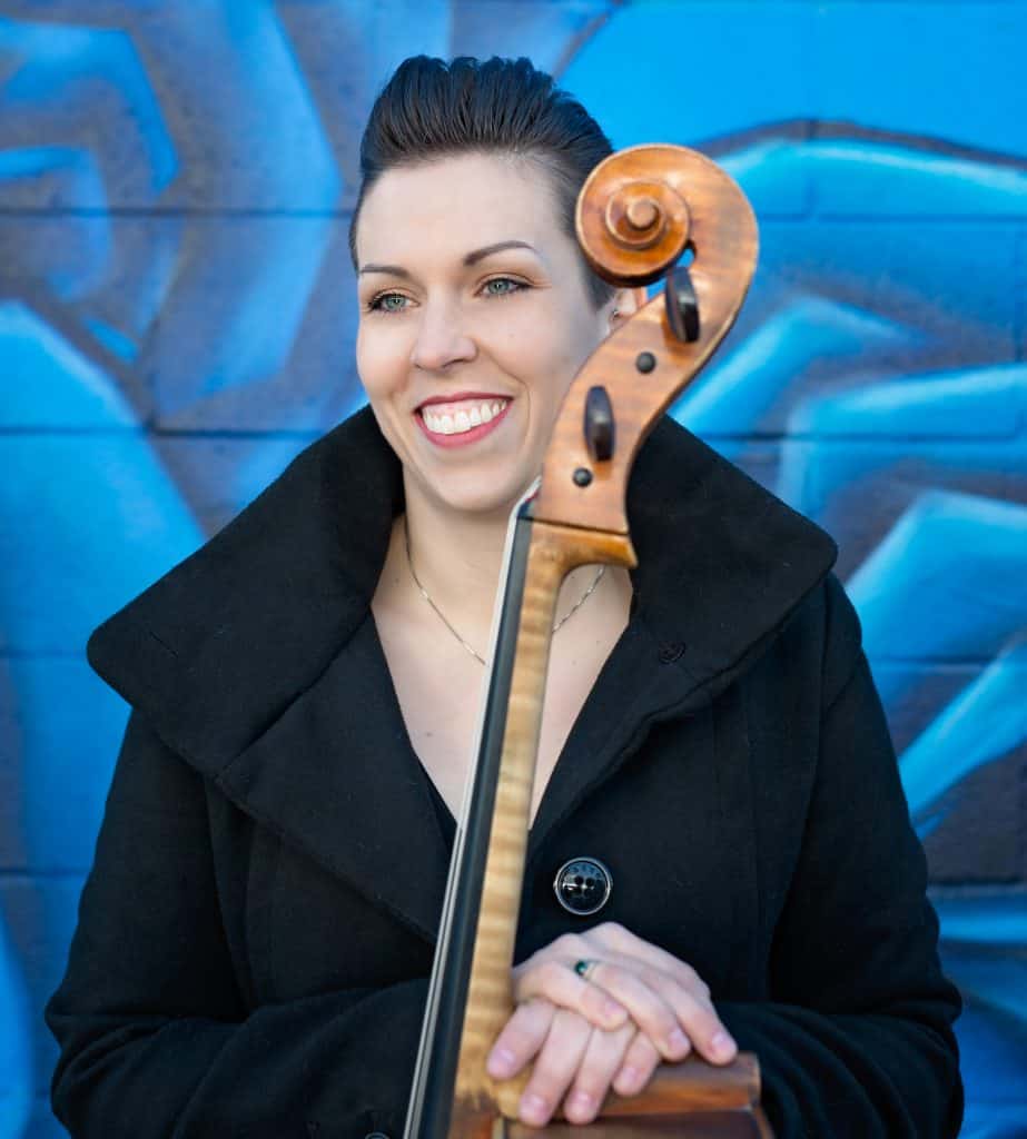 A woman in a black coat smiles while holding a double bass in front of a vibrant blue graffiti background.