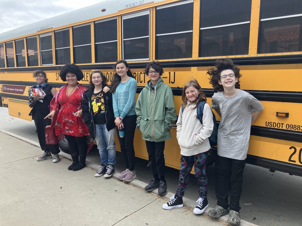 A diverse group of seven children stands smiling by a yellow school bus, with varying outfits and hairstyles.