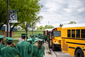 A group of graduates in green gowns walks towards yellow school buses under a partly cloudy sky.