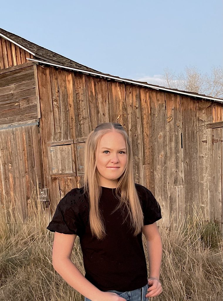 Breanne Baker, a Skyline High School Senior stands in front of a rustic wooden barn, with tall grass in the foreground.