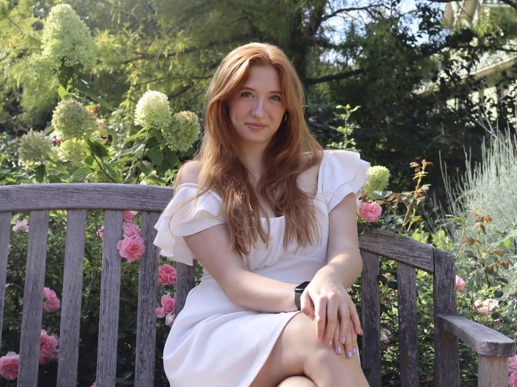 Katie Brownsberger, a Lyons Middle Senior High School Senior sits on a wooden bench surrounded by flowers in a sunny garden. She wears a white dress.