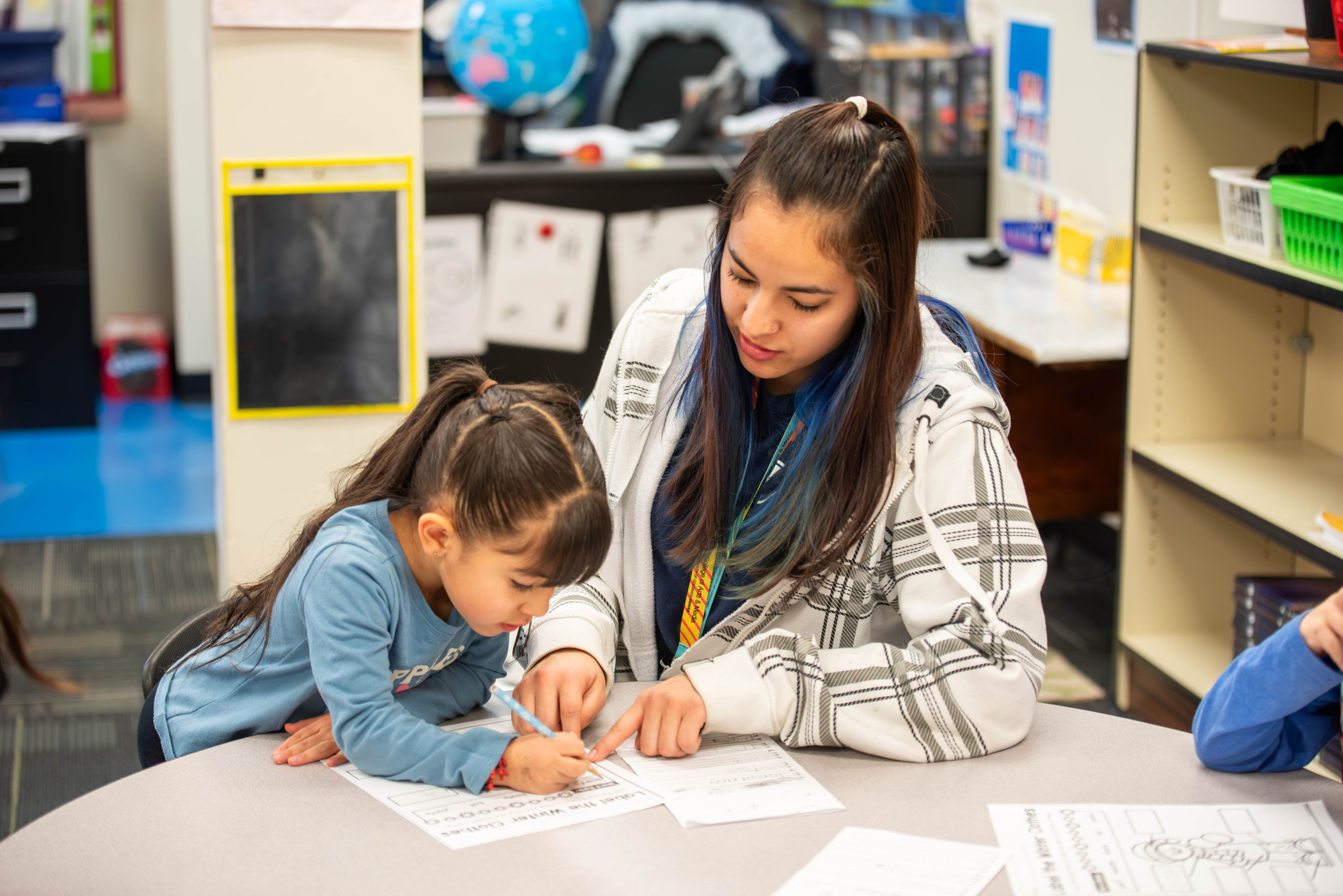 A young girl works on a worksheet with a P-TEACH mentor in a classroom setting. Educational materials are visible nearby.