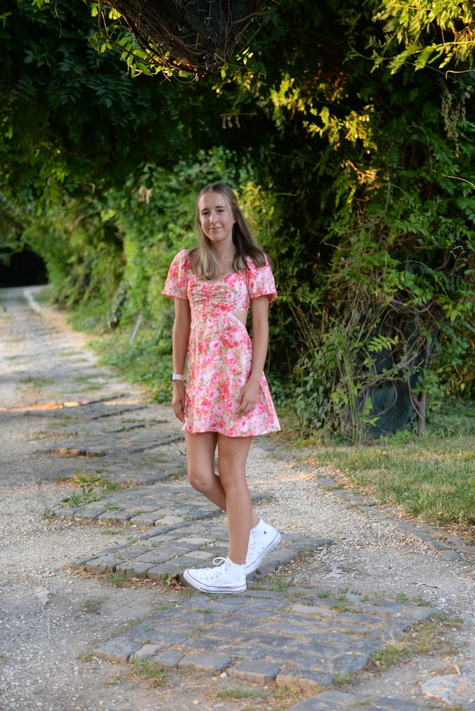 Sabina Stroia, an Eerie High Senior stands on a stone pathway, wearing a pink floral dress and white sneakers, surrounded by greenery.