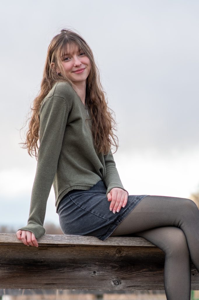 Eissa Everdeen, an APEX Homeschool Senior sitting on a wooden railing, dressed in a green sweater and gray skirt.