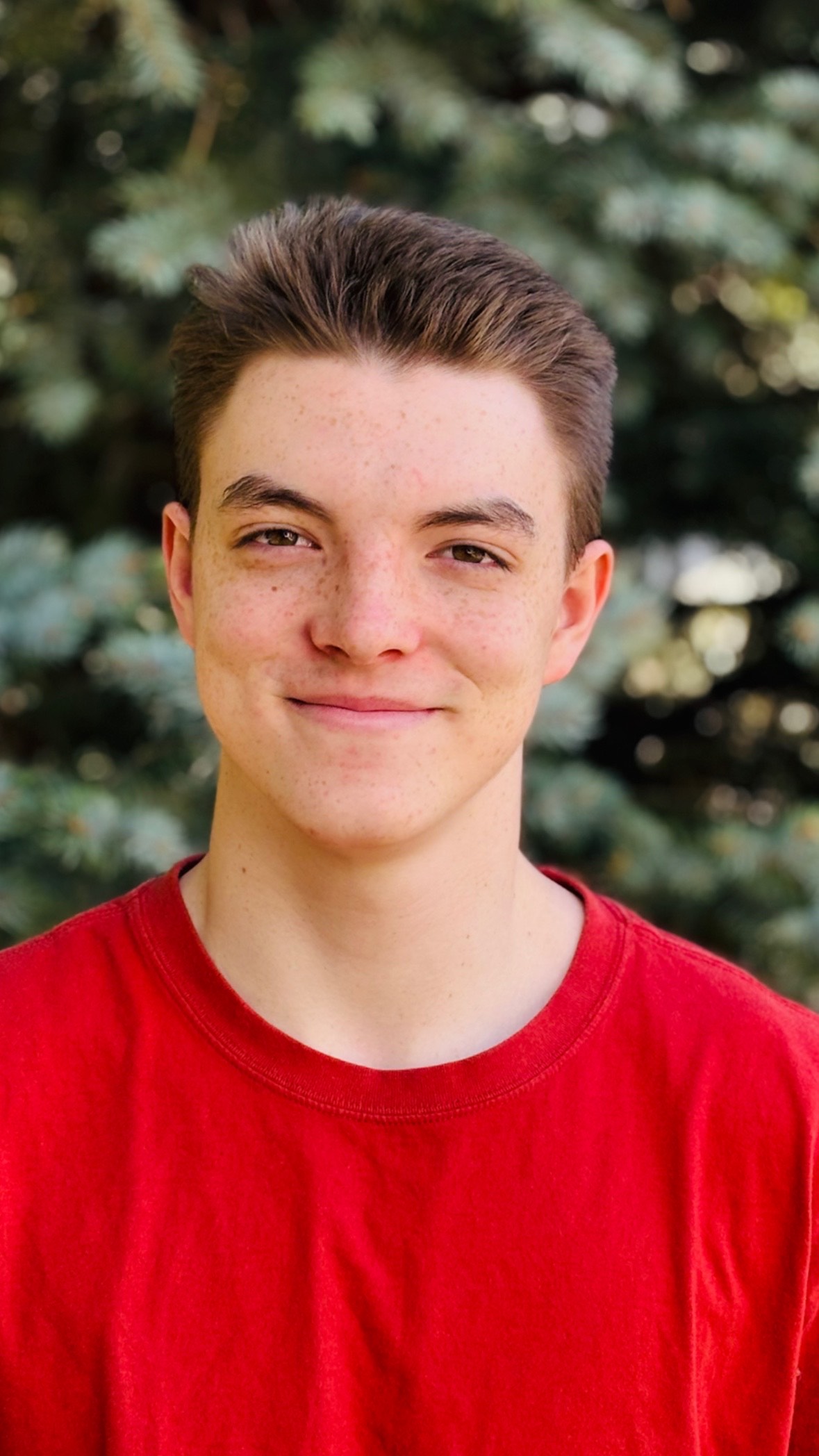 Luca Fowler, a St. Vrain Virtual High School Senior smiles, wearing a red shirt, with green foliage in the background.