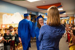 Graduates in blue caps and gowns celebrate in a school hallway while younger students applaud and cheer.