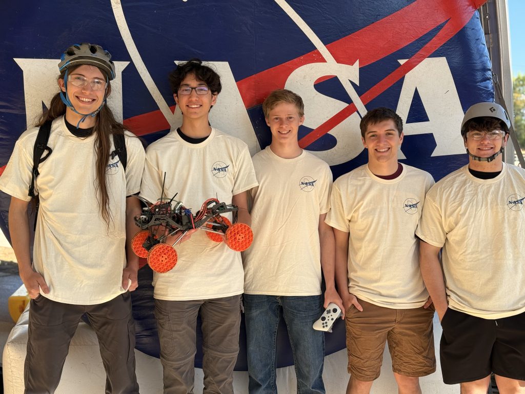 Cinco estudiantes con camisetas de la NASA posan juntos, sosteniendo un robot con ruedas, con una pancarta de la NASA al fondo.