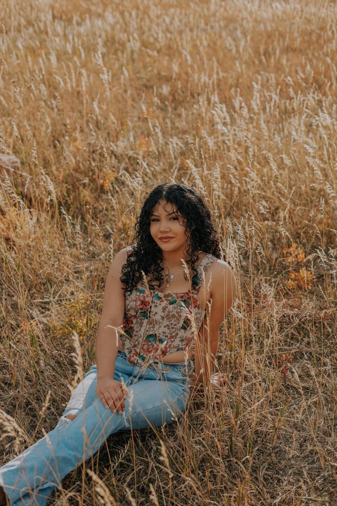 Karla Navarrete Espinoza, a Skyline High School Senior sits among tall golden grasses, wearing a floral top and light blue jeans.