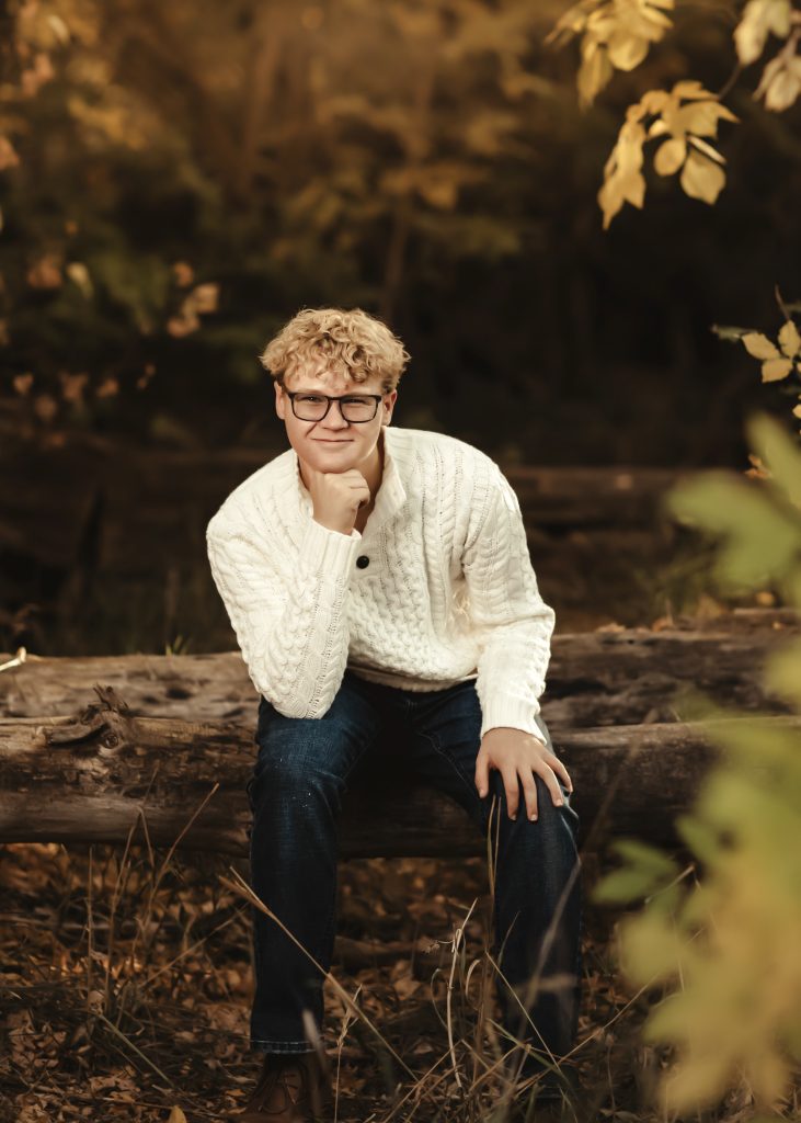 Colton Kominski, a Longmont High Senior sits on a log, wearing a white sweater, in a nature setting.