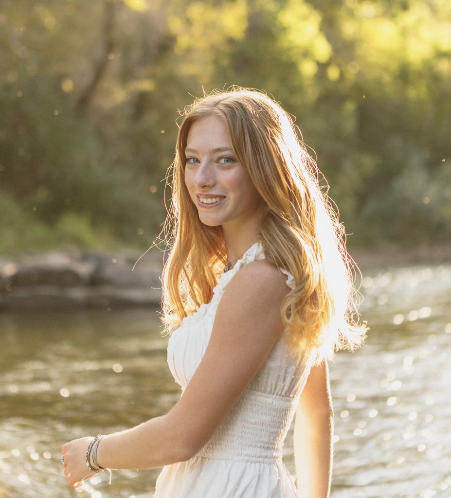 Clara Linder, a Longmont High Senior smiles by a river, sunlight illuminating her hair and the surrounding trees.