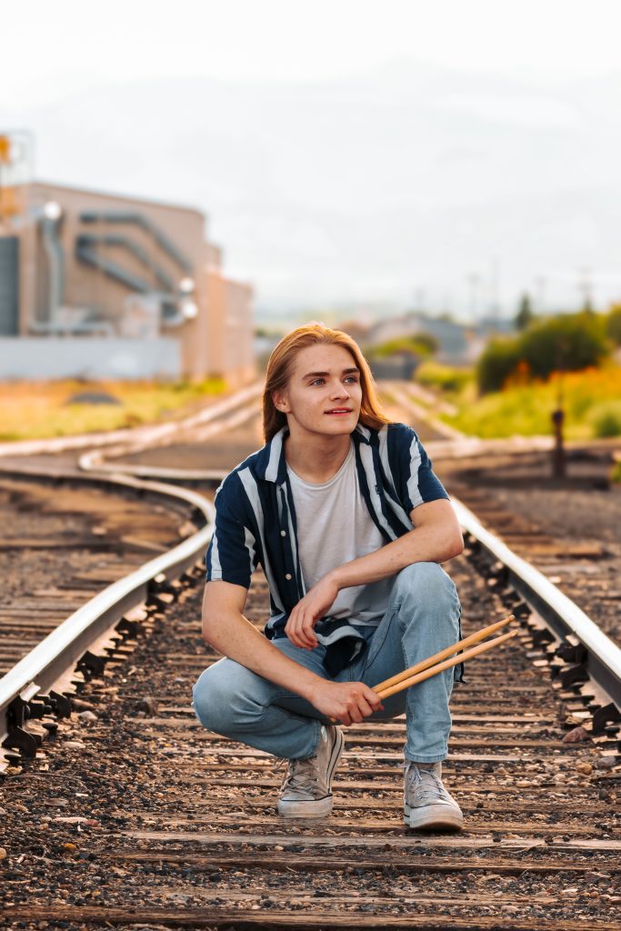TJ Mogensen, a Frederick High Senior crouches on train tracks, holding drumsticks, with a background of greenery and buildings.