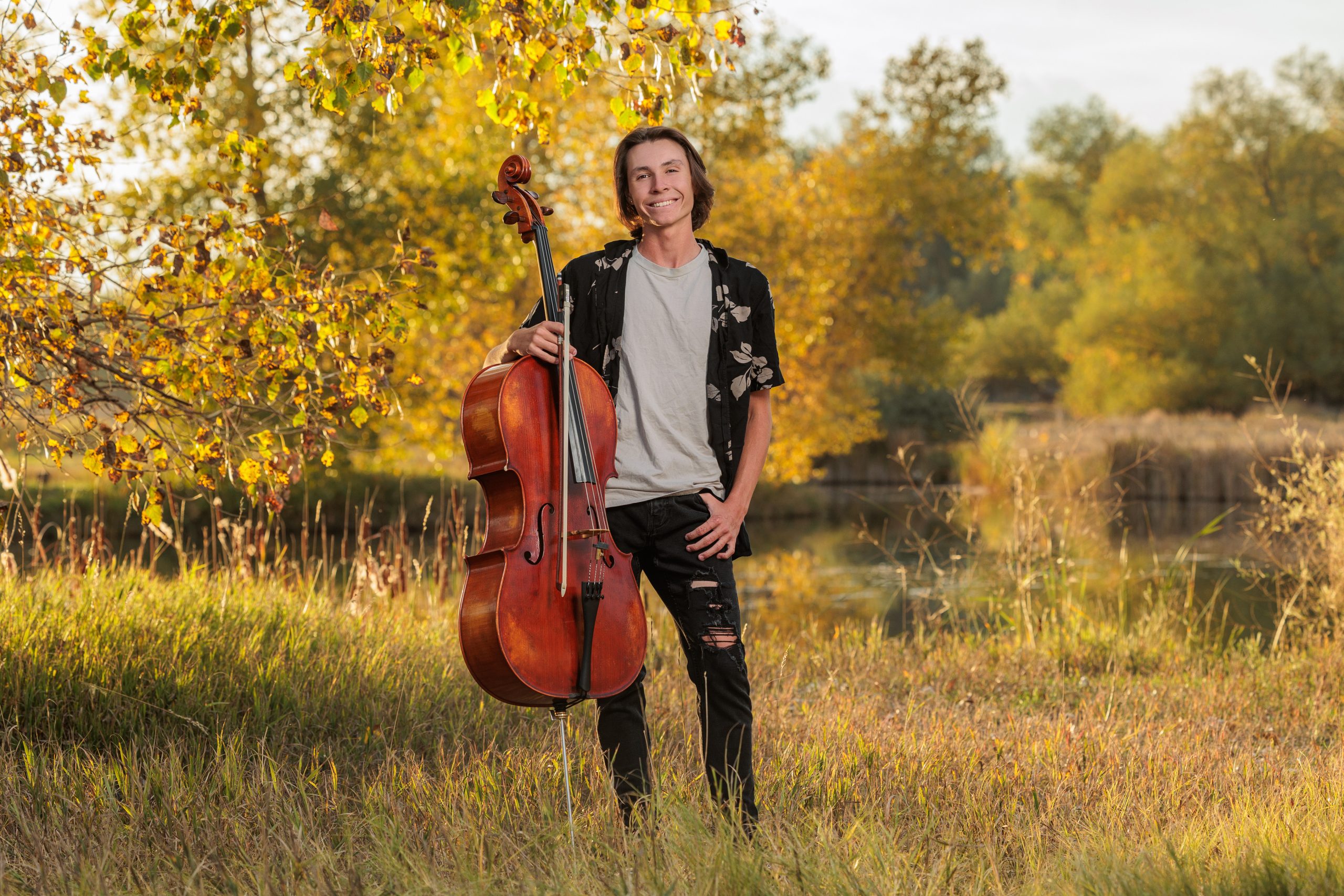Atlas Momier, an Erie High Senior in a field with a cello, surrounded by autumn foliage and a calm water backdrop.
