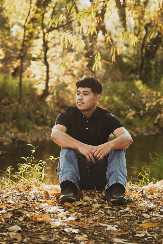 Bryan Perdomo Deleon, a Frederick High Senior sits on autumn leaves near a pond, surrounded by trees with golden foliage, looking contemplative.