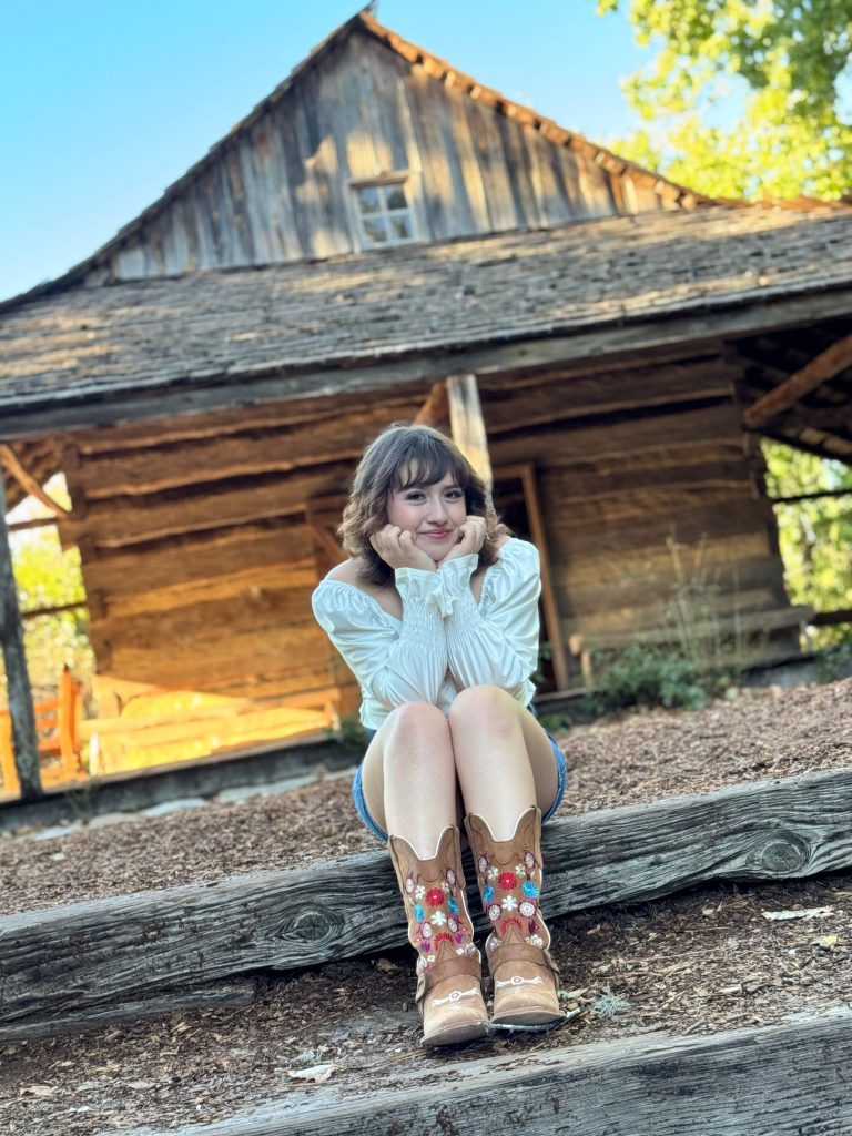 Erika Rubio, a LaunchED Senior sits on wooden steps, smiling with an old cabin in the background.