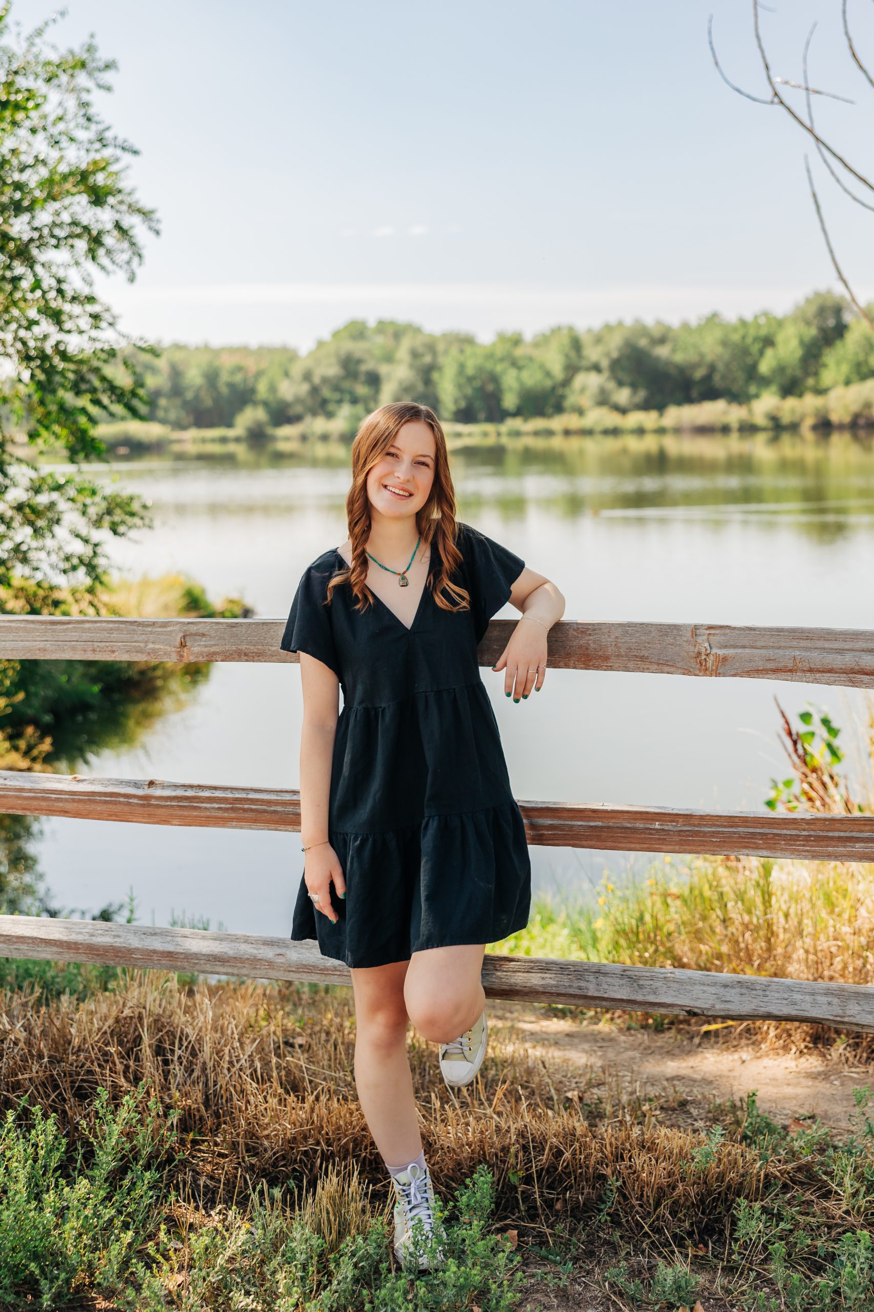 Paige Schleper, a Silver Creek High School Senior leans on a wooden fence by a serene lake, surrounded by greenery.