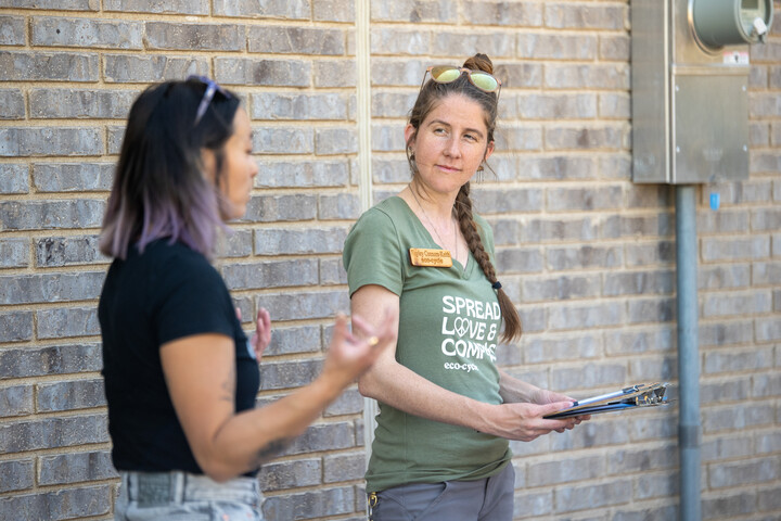 Two women are engaged in conversation outside by a brick wall; one holds a clipboard and wears a name tag.