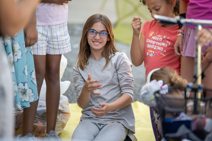 A girl with glasses smiles while sitting on the ground among other children, engaged in a playful activity.