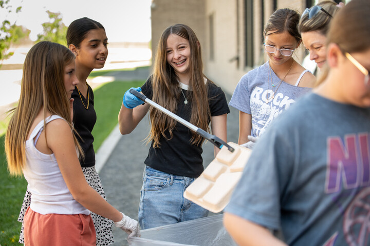 A group of young people smiles while participating in a cleanup activity, holding tools and a trash bag outdoors.