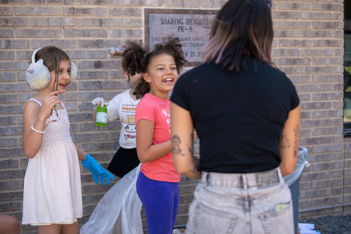 Children smiling and engaging in an outdoor activity, with one adult guiding them near a school building.