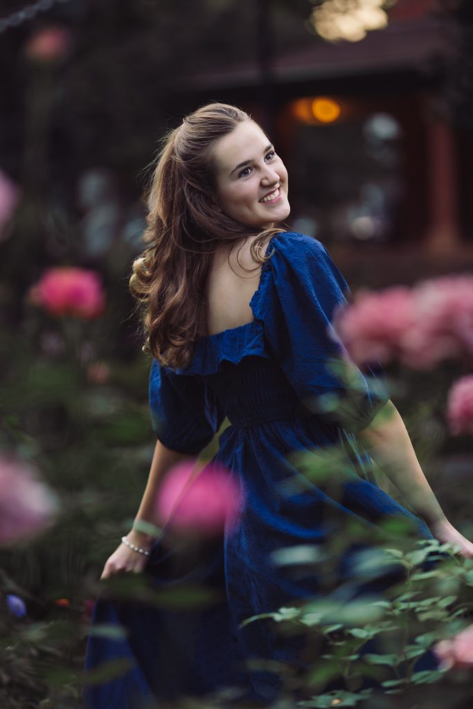 Raelan Schmidgall, an APEX Homeschool Senior smiles over her shoulder among pink flowers in a garden.