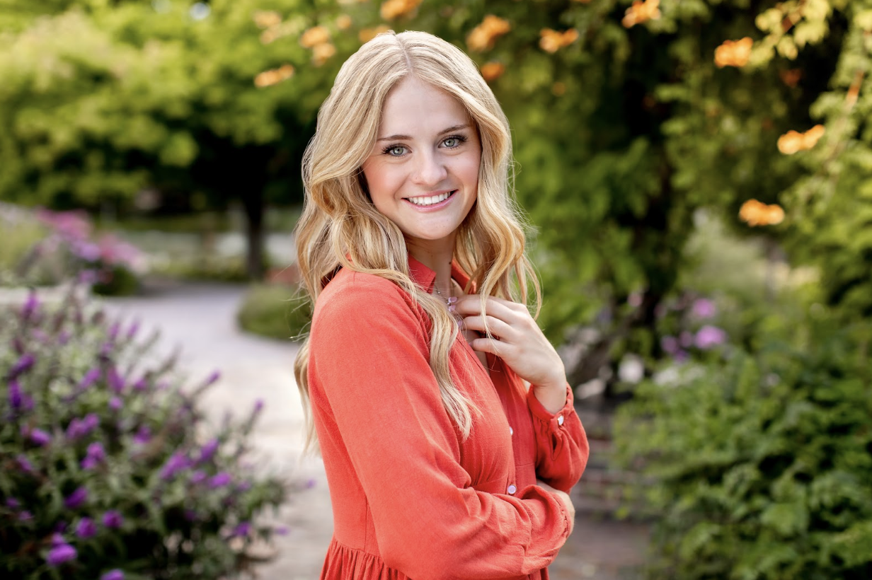 Adisyn Rademacher, a senior at Mead High School stands among lush greenery and flowers, with a pathway in the background.