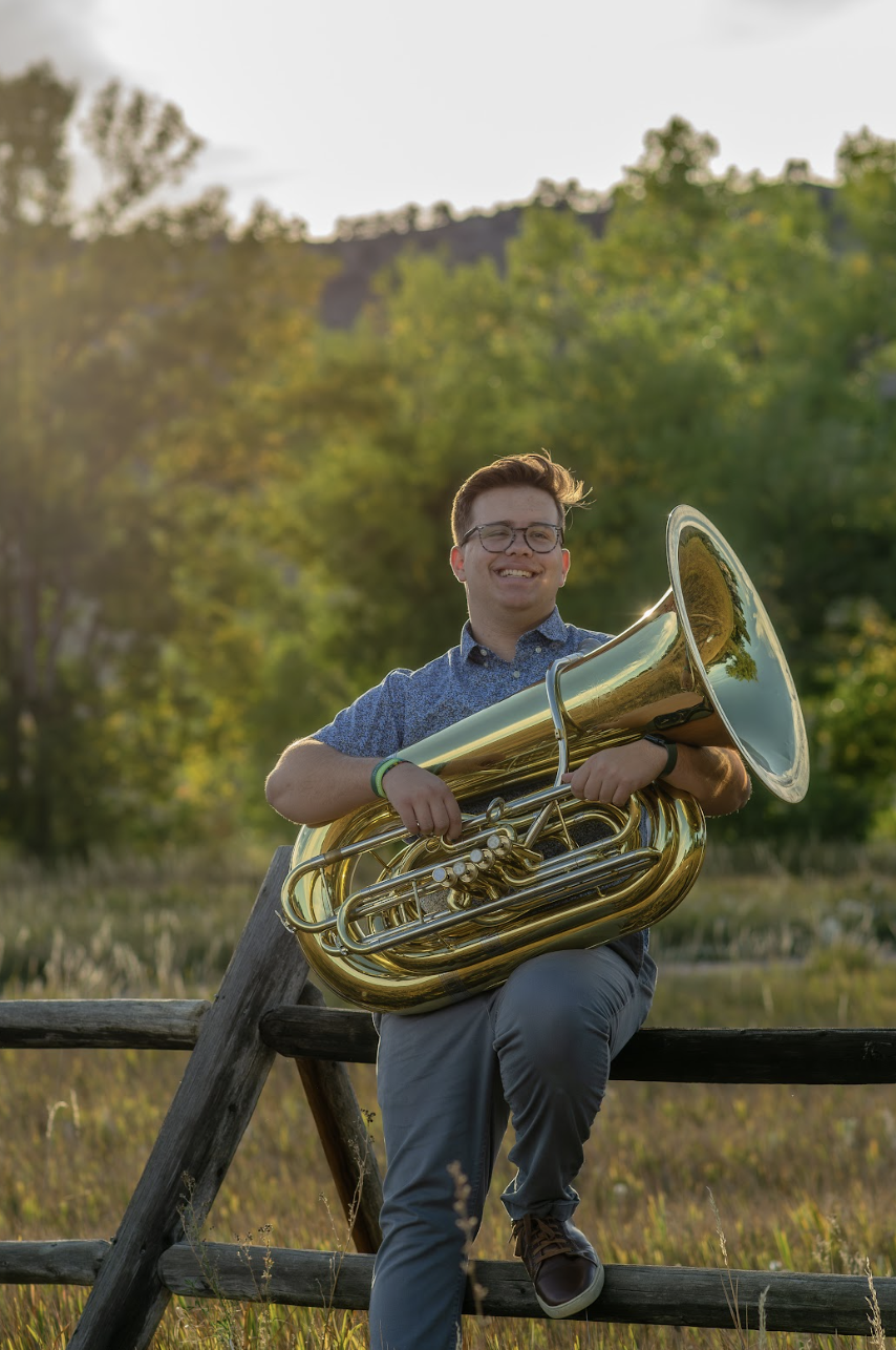 Zachary Kaufman, a Silver Creek High School Senior sits on a wooden fence holding a tuba, surrounded by greenery in a sunny outdoor setting.