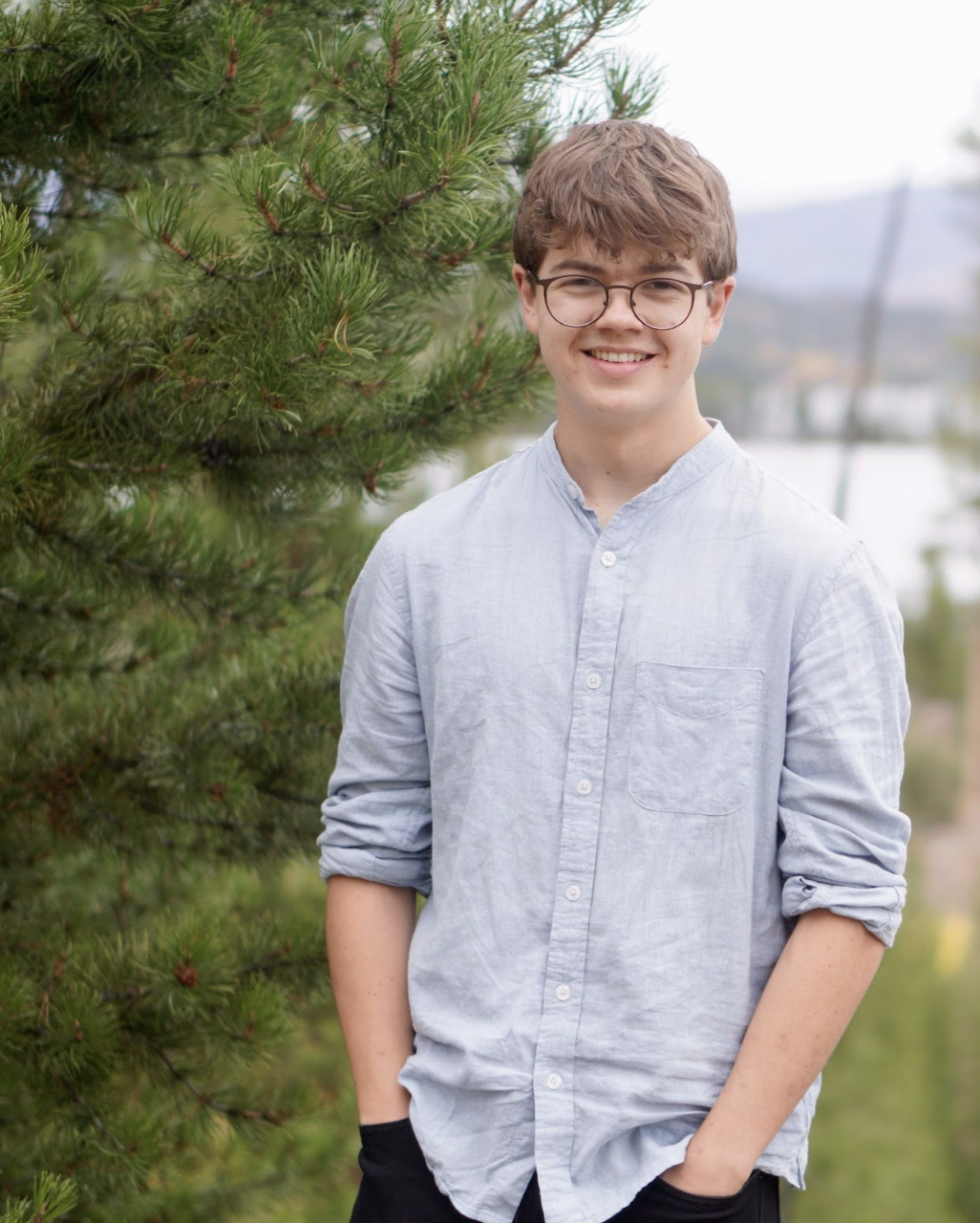 Jack McGuirk, a Niwot High School Senior stands by a pine tree, wearing a light blue shirt and black pants, with a scenic background.