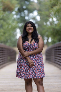 A person with long black hair smiles while wearing a floral dress, standing on a wooden bridge surrounded by greenery.