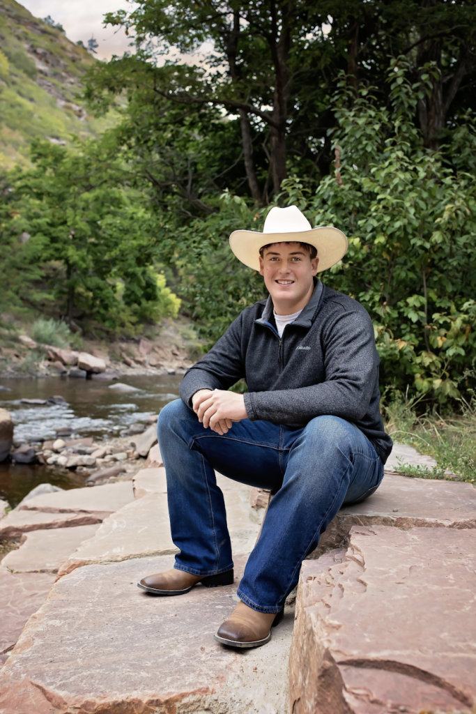 Grant Gordon, a Mead High School Senior sits on rocks by a stream, surrounded by greenery, wearing casual clothes and boots.