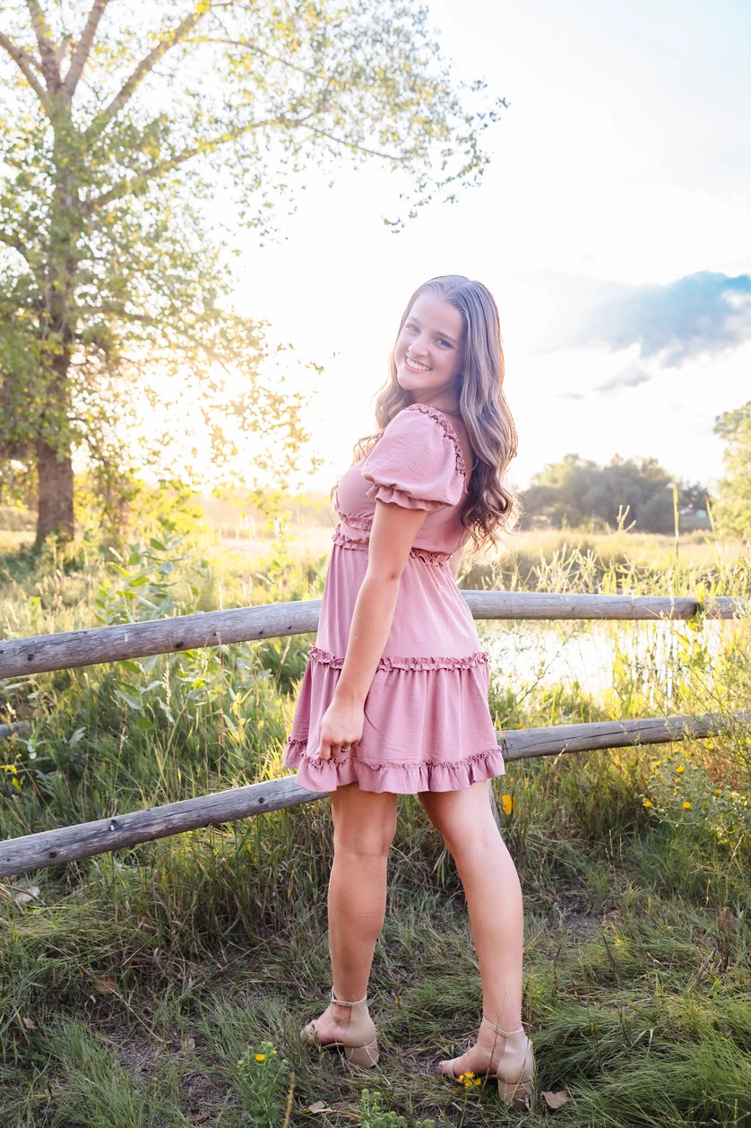 Zoe Wittler, a Frederick High Senior smiles while standing near a fence, with a sunset backdrop and greenery surrounding her.