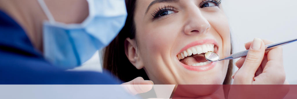 A smiling woman sits in a dental chair as a dentist examines her teeth with a dental tool.