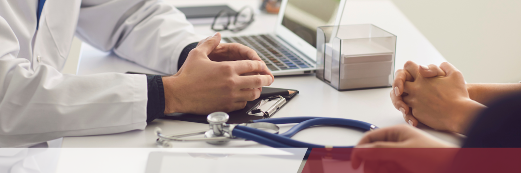 Se ven las manos de un médico mientras habla con un paciente; sobre la mesa hay un ordenador portátil y instrumental médico.