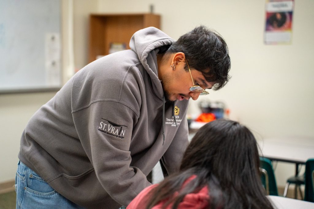 A person in a hoodie leans over a table to assist another student in a classroom setting.