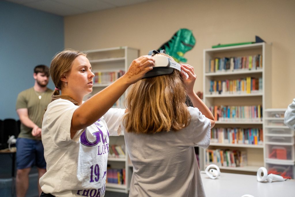 Two people are in a room, one is fitting a virtual reality headset on another, while a third person observes.