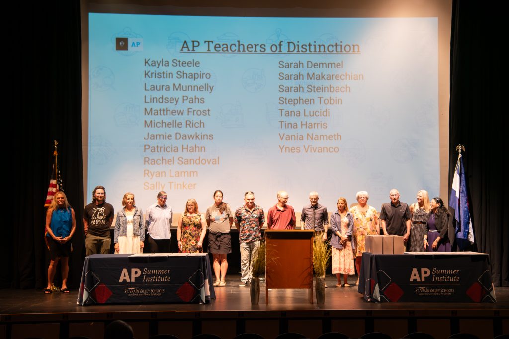 Un grupo de profesores posa en el escenario durante un acto en homenaje a los profesores destacados del programa AP, con una presentación a sus espaldas.