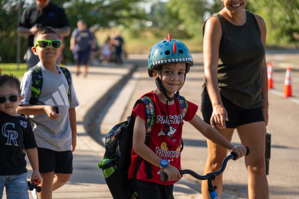 Un niño con un casco de colores va en patinete, sonriendo mientras pasea con sus amigos y unos adultos en un día soleado.