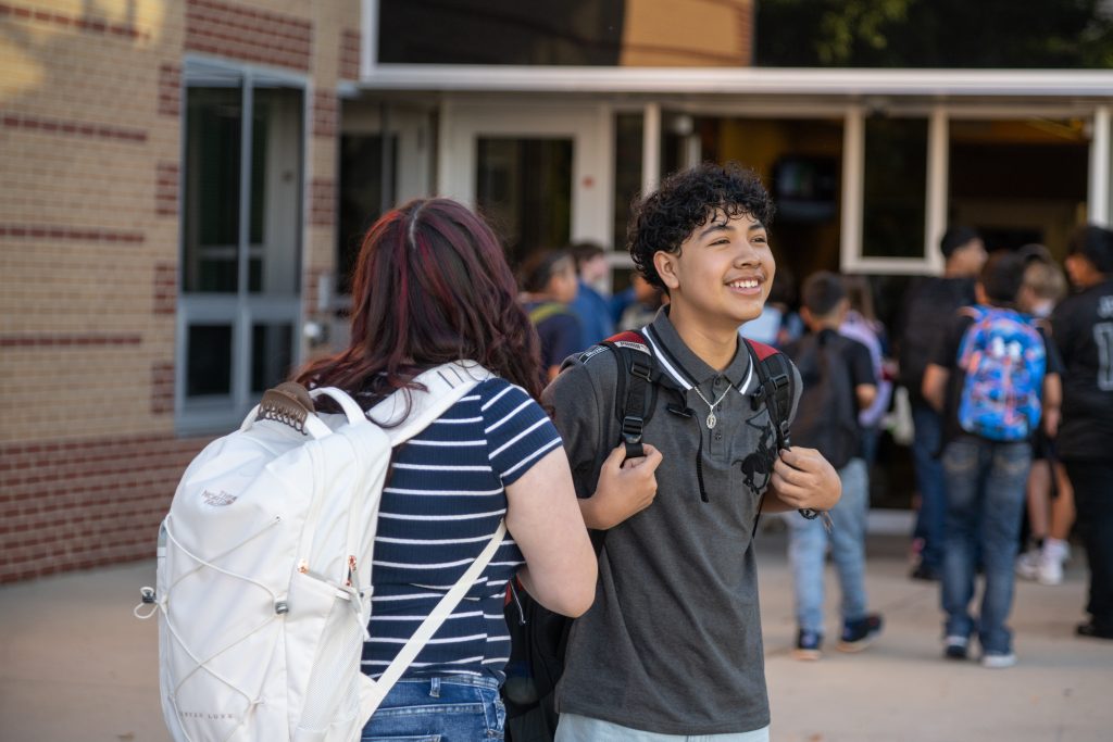 Unos alumnos charlando a la salida de un colegio; uno de ellos sonríe y el otro lleva una camiseta a rayas y una mochila.