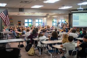 Un aula llena de alumnos que levantan la mano, concentrados en una clase impartida por un profesor al frente.